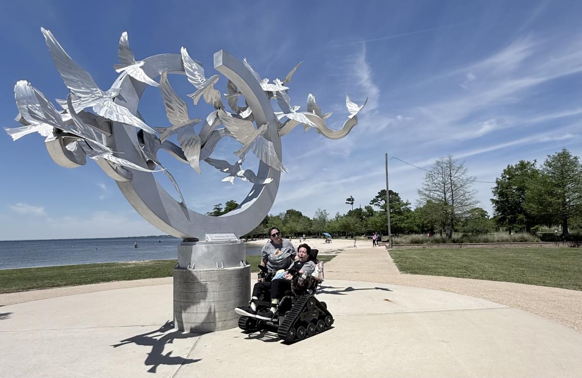 Christy and PJ Paulsell pause beside Bird's Eye View, a soaring metal sculpture by Aly Adduci with Gabriel Wimmer and Brent Barnidge, while PJ rides in an all-terrain TrackChair at Fontainebleau State Park overlooking Lake Pontchartrain.