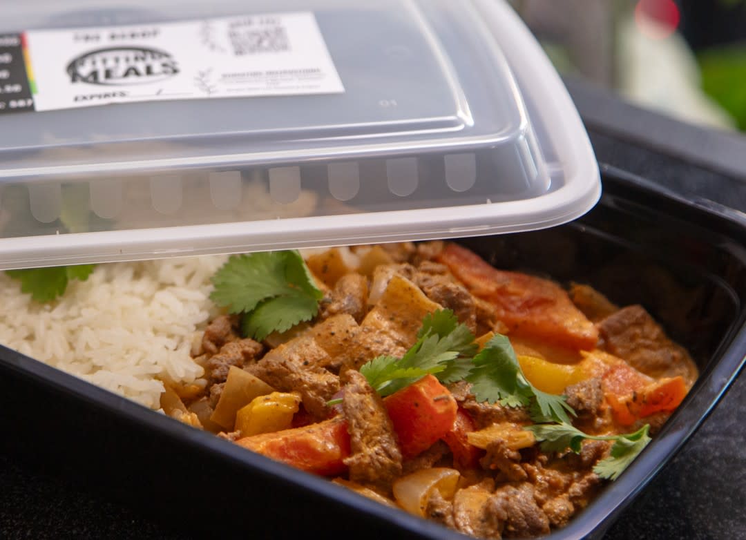 A black meal container with partly opened lid reveals white rice and colorful beef stir-fry with bell peppers and cilantro.