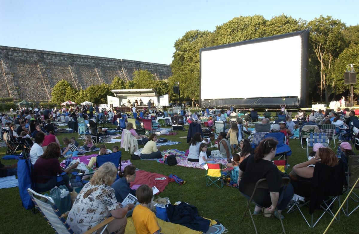 A large gathering of people sit on picnic blankets and chairs facing toward a giant screen.