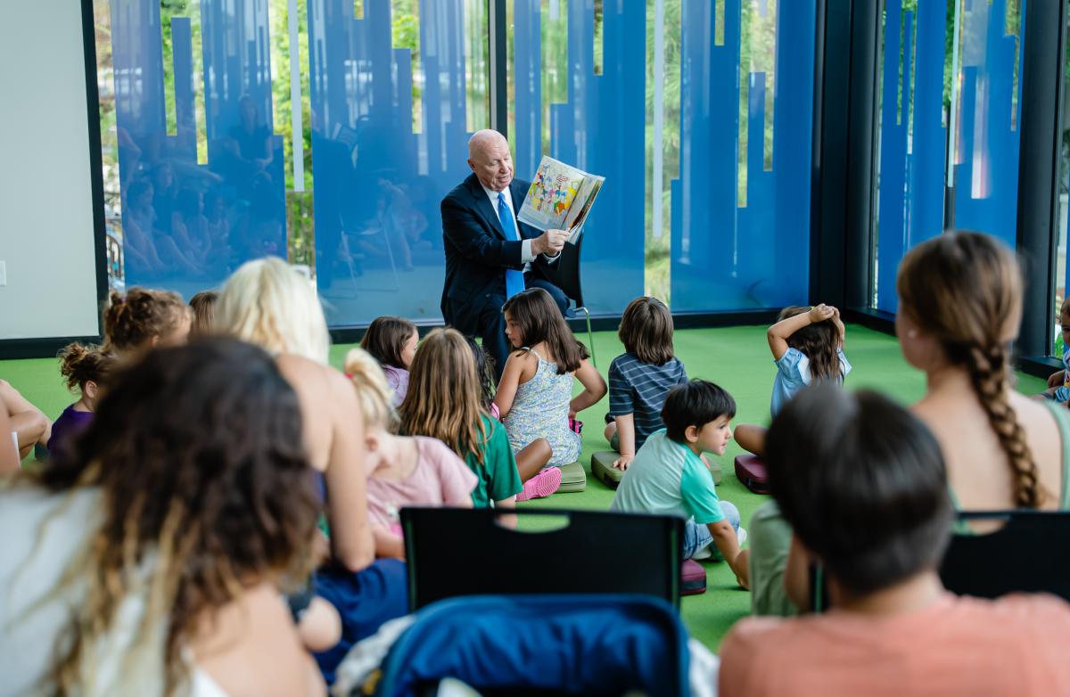 During the grand opening of the library, Kevin Brady reads the children's book, "House Mouse, Senate Mouse." About a dozen children sit on the floor in front of him (their backs to the camera). The camera peeks through the heads of parents sitting behind them.