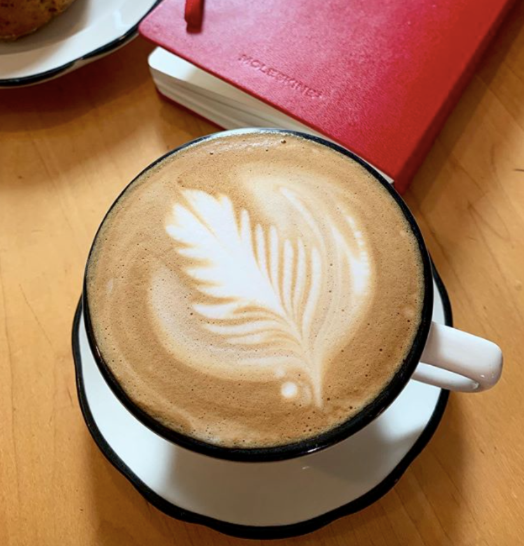 A white coffee cup on a white plate sits on a wooden table. The coffee has a design on top made from cream.