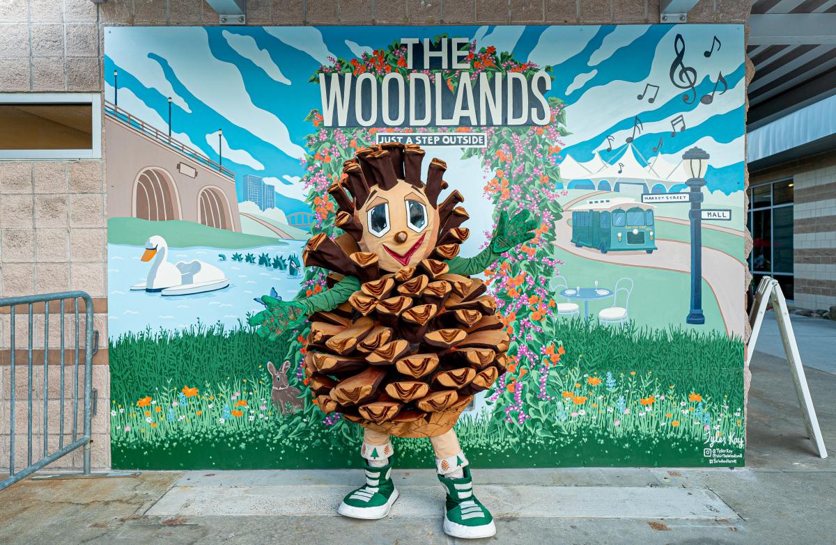 Puffy The Pinecone Poses in front of Visit The Woodlands Mural