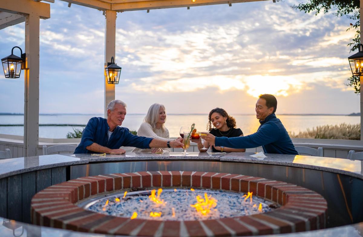 Four people gather around a circular fire pit, toasting drinks as the sun sets over a serene waterfront. Lanterns illuminate the scene.