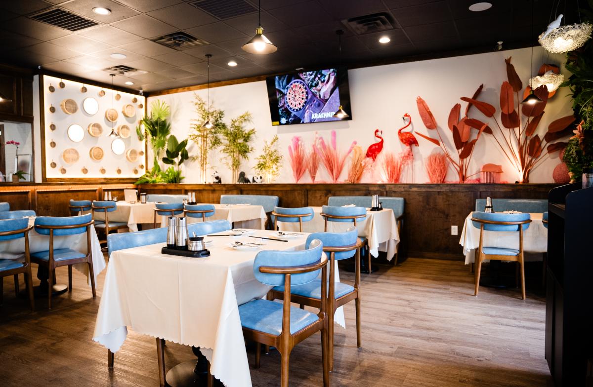 An overview of the Mimi Garden dining room. The frame includes six cloth-covered tables with light blue chairs. The back wall is decorated with a TV and fake but tasteful, colorful foliage. The right half features pink tropical plants and flamingos, and on the left, bamboo stalks and pandas. A dividing wall on the left displays woven dumpling baskets in various sizes.