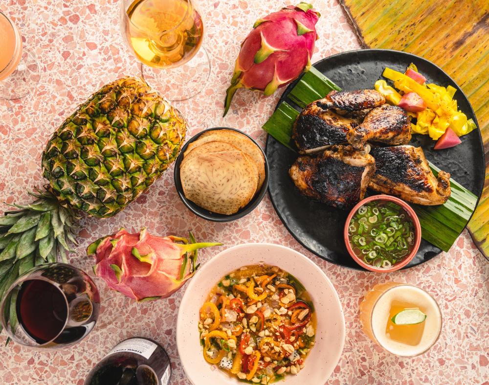 Spread of colorful Carribean food on neutral plates. Jerk chicken and roti sit alongside whole dragon fruit and pineapple.