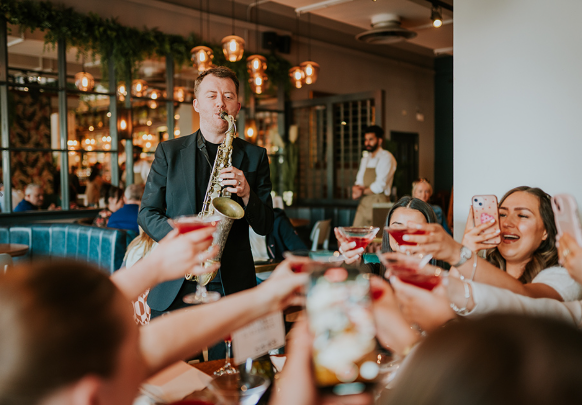 Saxophone player playing to group of women toasting their cocktail glass at All Bar One