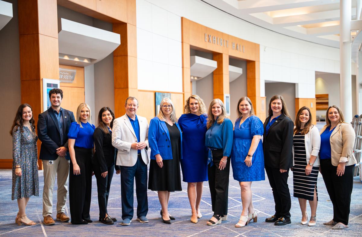 Twelve people smile at the camera before the Exhibit Hall at The Woodlands Waterway Marriott Hotel & Convention Center. Everyone is dressed professionally and all wearing something in bright royal blue in honor of GMID. From left to right: Andrea Day, Ryan Greenwood, Brooklyn Newell, Carla Twidell, Nick Wolda, Julie Quinn, Josie Lewis, Ashley Fenner, Elizabeth Eddins, Rachael Varner, Ashley White, and Amber Parsley.