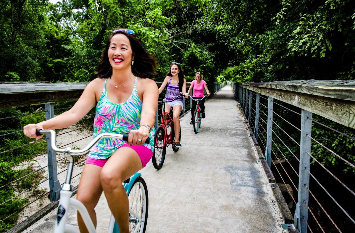 Three cyclists ride across a bridge on the Tammany Trace, surrounded by lush green trees along the scenic rail trail on a summer day.