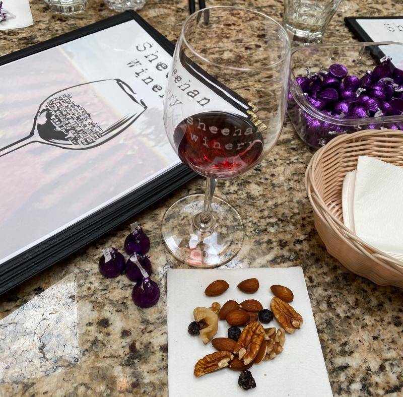 A Sheehan Winery menu, a glass of wine, some chocolate and some nuts sit on a table.