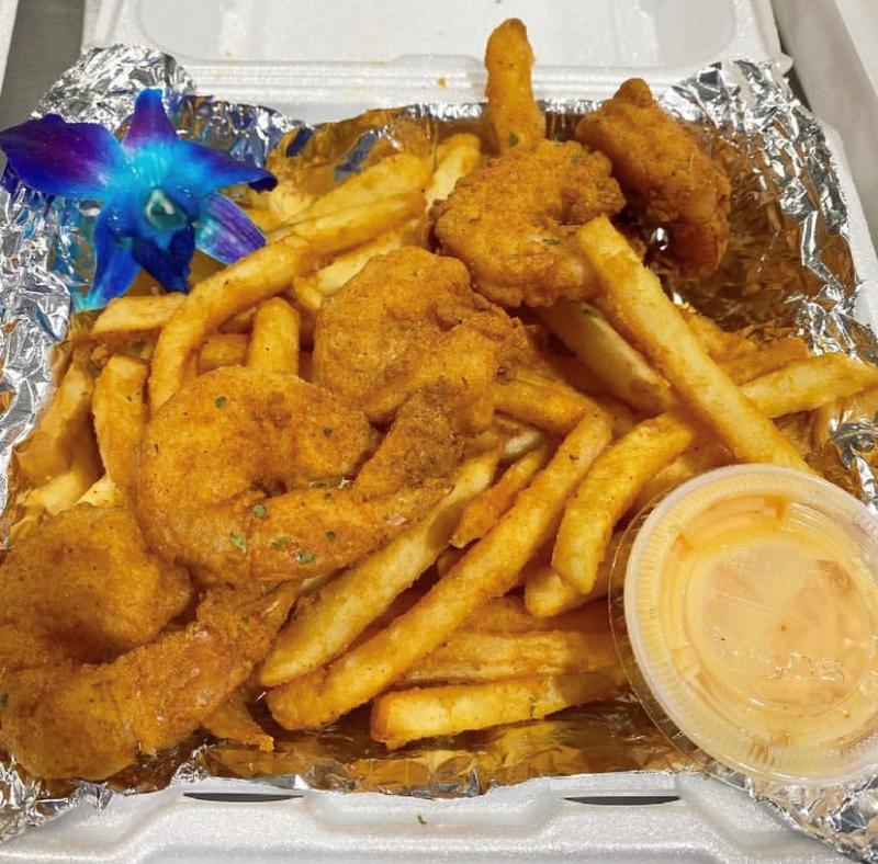 fried shrimp overtop fries in a to go tray with tin foil on the bottom featuring a blue flower garnish