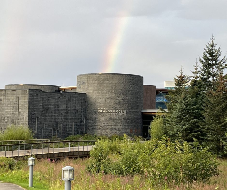 he Alaska Islands & Ocean Visitor Center with a soft rainbow arching over it, surrounded by lush green trees and shrubs, on a partly cloudy day.