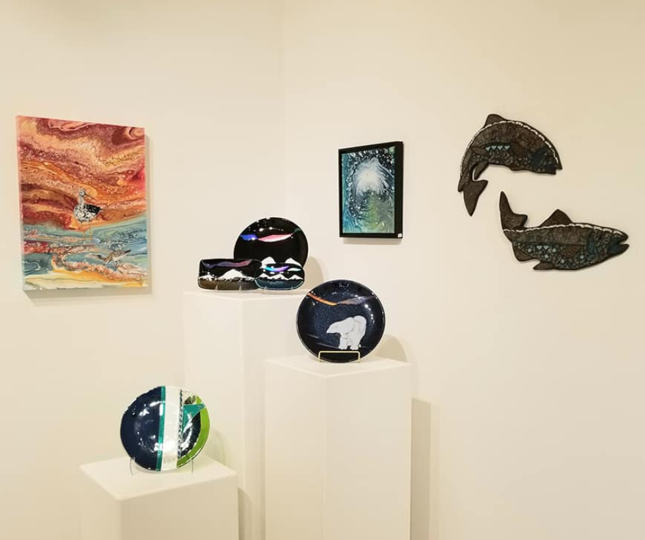 A collection of vibrant and contemporary Alaskan art pieces, including colorful glass bowls, abstract paintings, and whale wall art displayed on white pedestals and walls.