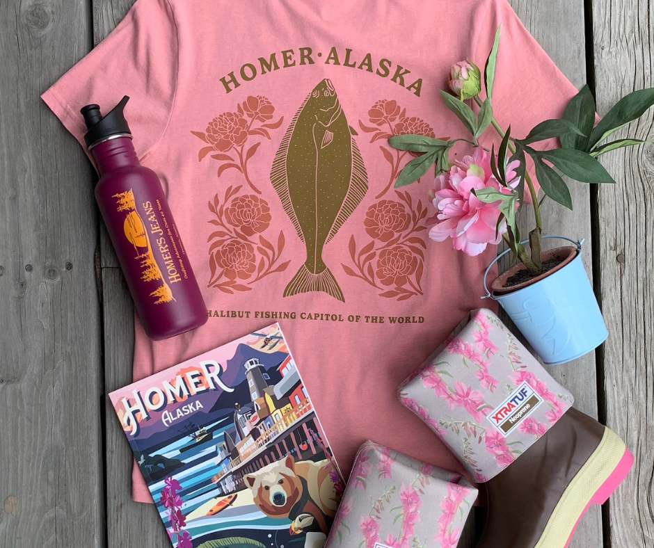 A spread of Homer-themed merchandise including a pink t-shirt with a halibut illustration, a maroon water bottle, a Homer travel guide, and pink floral XtraTuf boots, arranged on a wooden surface.