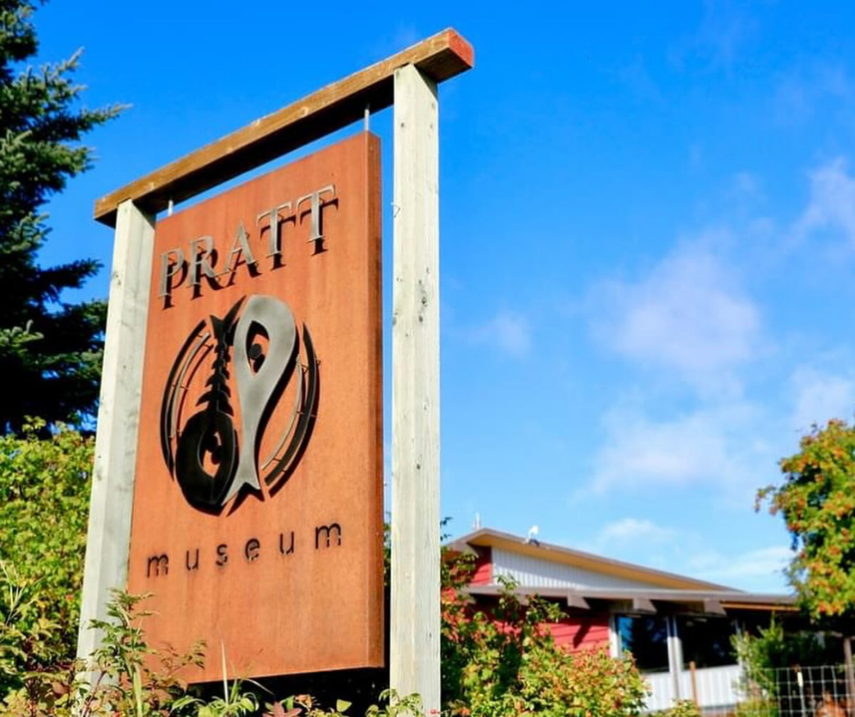 The sign for the Pratt Museum in Homer, featuring a fish and feather logo against a bright blue sky, with part of the museum building visible in the background.