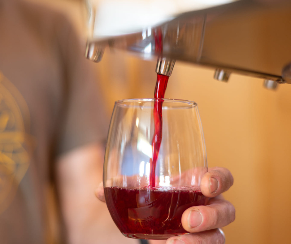 A close-up shot of a hand pouring a rich, deep red mead into a glass, highlighting the color and texture of the beverage.