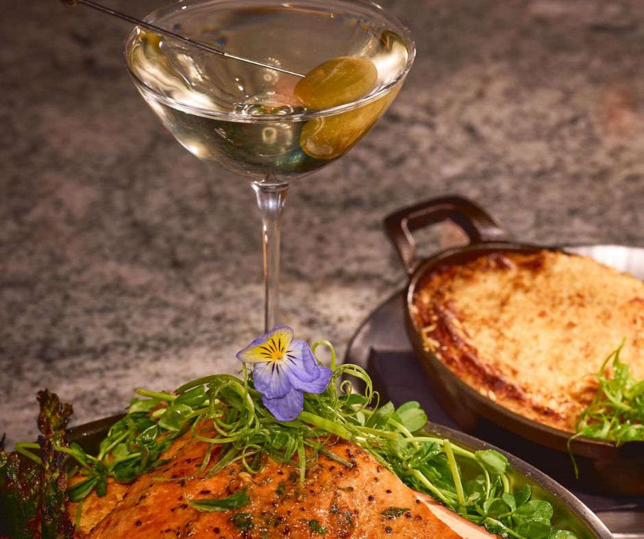 A gourmet meal with a beautifully plated salmon dish garnished with greens and an edible flower, accompanied by a martini and a dish of au gratin potatoes.