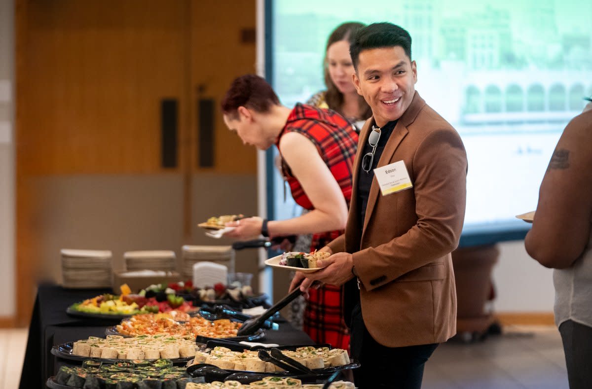 A man smiles at a person out of frame as he adds food to his plate at a buffet line at a meeting.