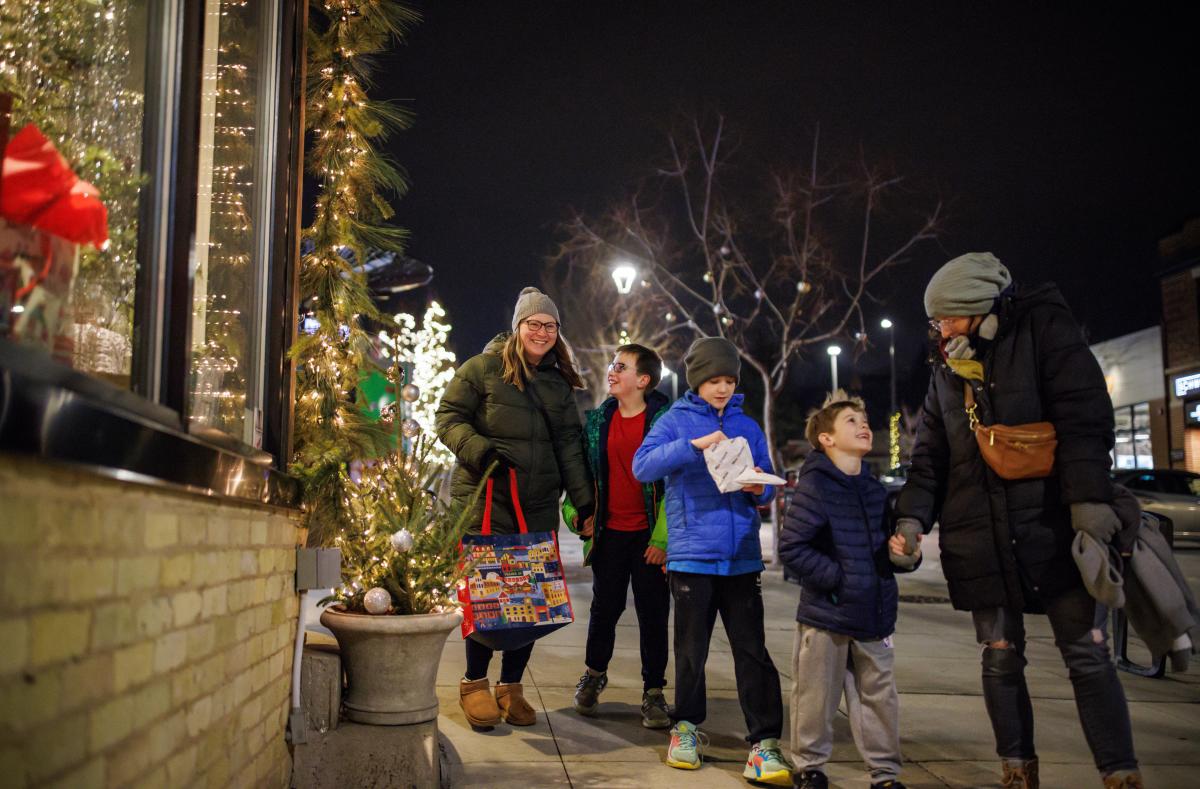 A group of two adults and three children walk together on a decorated city sidewalk at night during the holiday season. They are bundled in winter coats, hats, and gloves. Festive lights and small decorated trees line the shop windows on the left. One adult carries a colorful tote bag, and one child holds a snack. Everyone appears cheerful and engaged with one another as they enjoy the winter evening.”