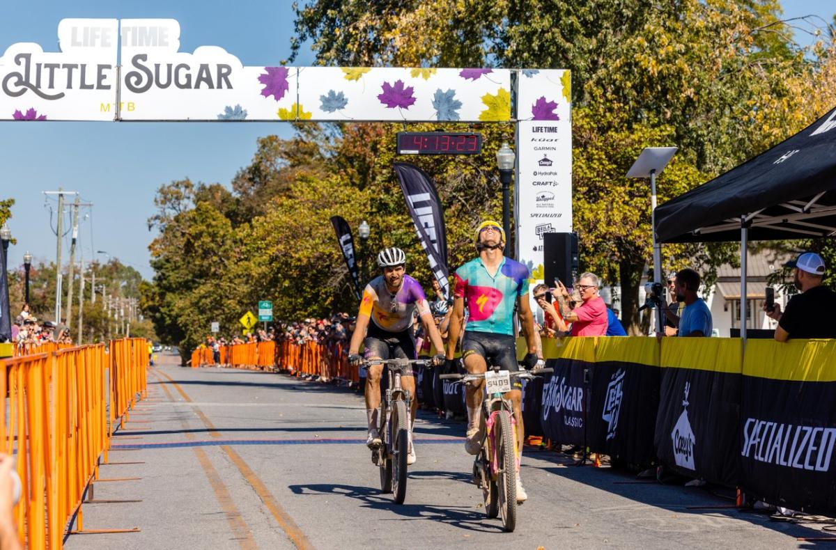 Two cyclists cross the finish line under a colorful arch at a biking event, surrounded by cheering spectators and vibrant autumn foliage.