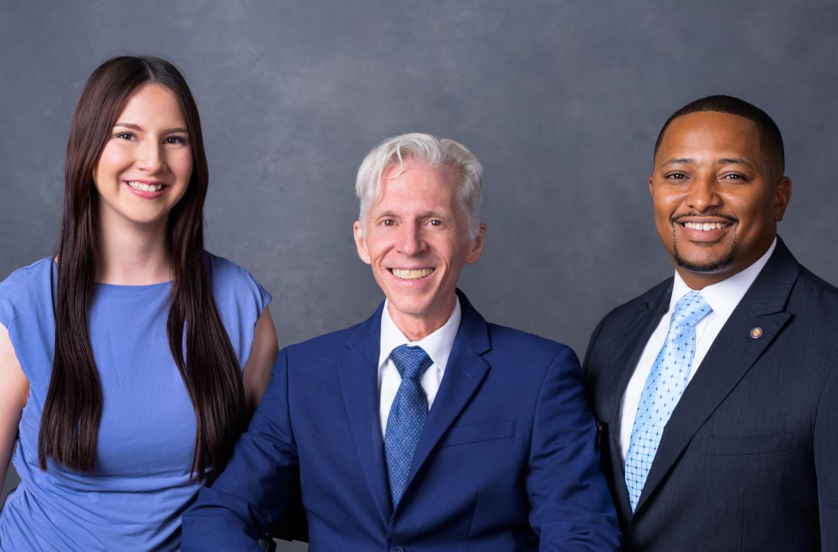 A diverse group of three smiling professionals dressed in formal business attire, with two men in suits and a woman in a light blue dress, against a gray background.