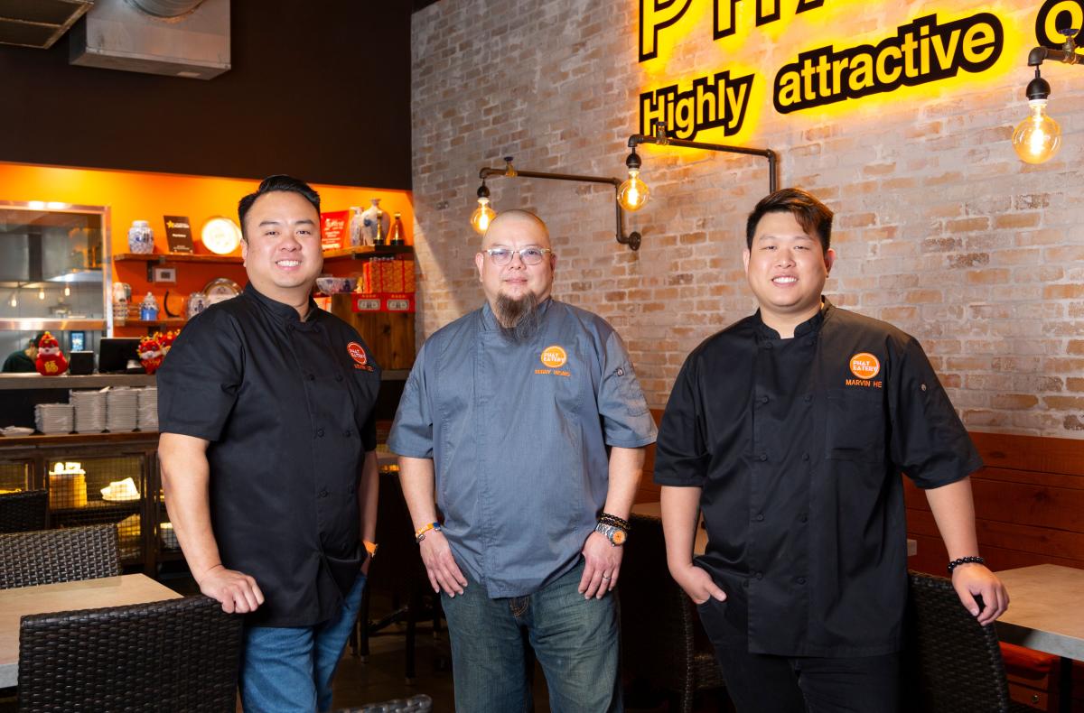 The camera faces Marvin He, Terry Wong, and Kevin Lee under the neon yellow "Phat (adjective) Highly attractive or gratifying" sign inside Phat Eatery.