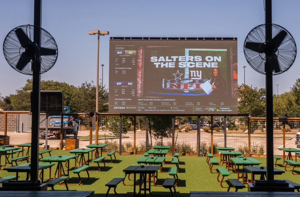 The Kirby Ice House Patio features dozens of green picnic tables, some shaded by the building, with large fans overhead and a giant projector screen showing sports stats in the back.