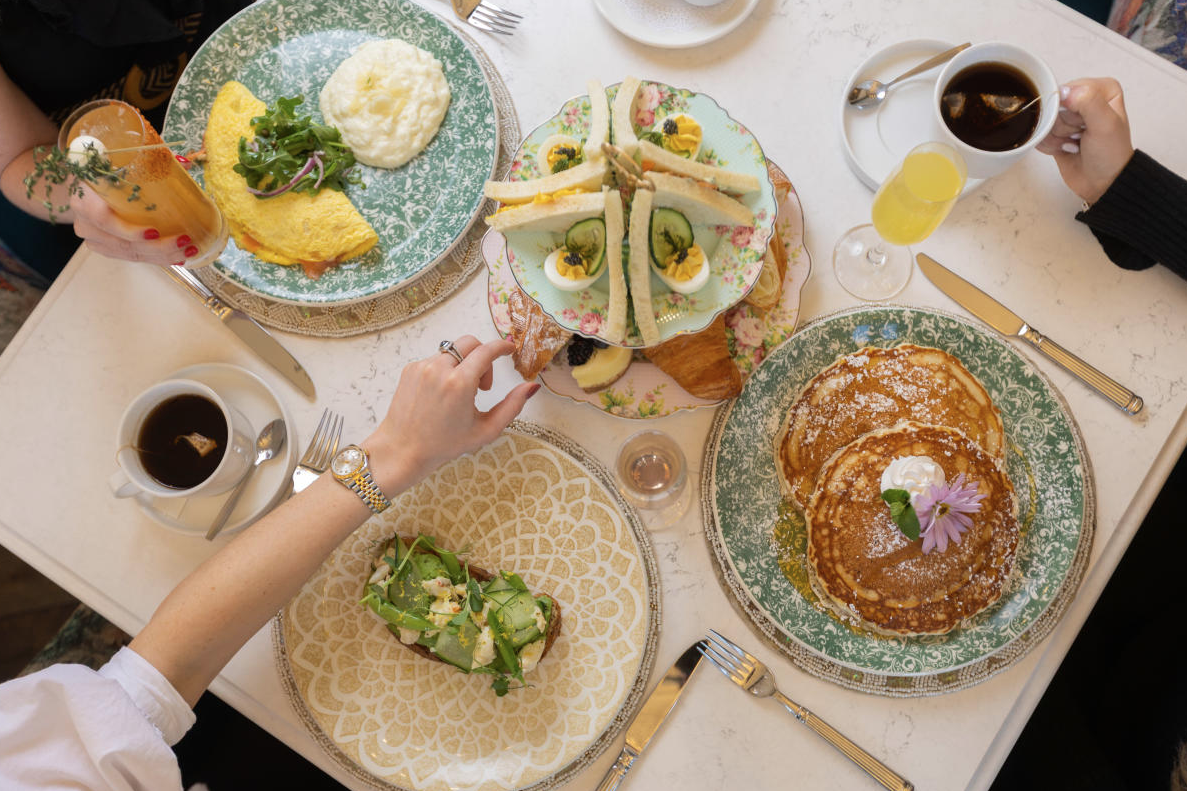 An overhead shot of brunch at The Audrey. The table is set for three with decorative pastel plates--one with an omelet and grits, one with avocado toast, one with fluffy pancakes--and a two-tiered tower of finger sandwiches, croissants, and deviled eggs. There're also two coffees and two mimosas.