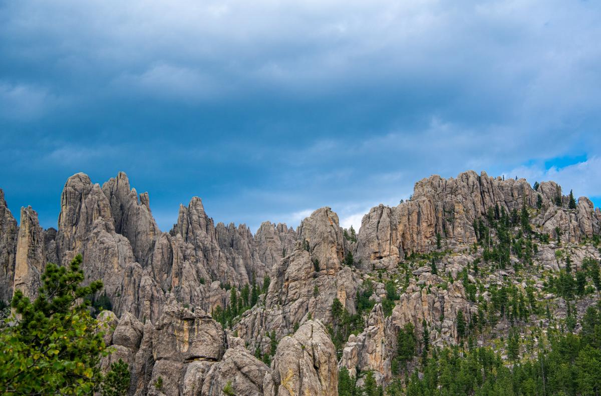 Rocky peaks under a moody blue sky, with green pine trees dotting the rugged terrain. The scene conveys a sense of vastness and solitude.