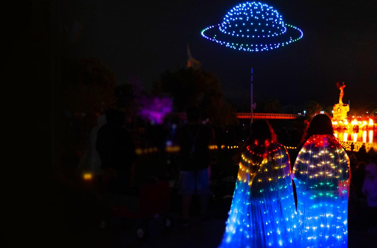 Two people adorned in led costumes watch as drones create a saturn over the Keeper of the Plains