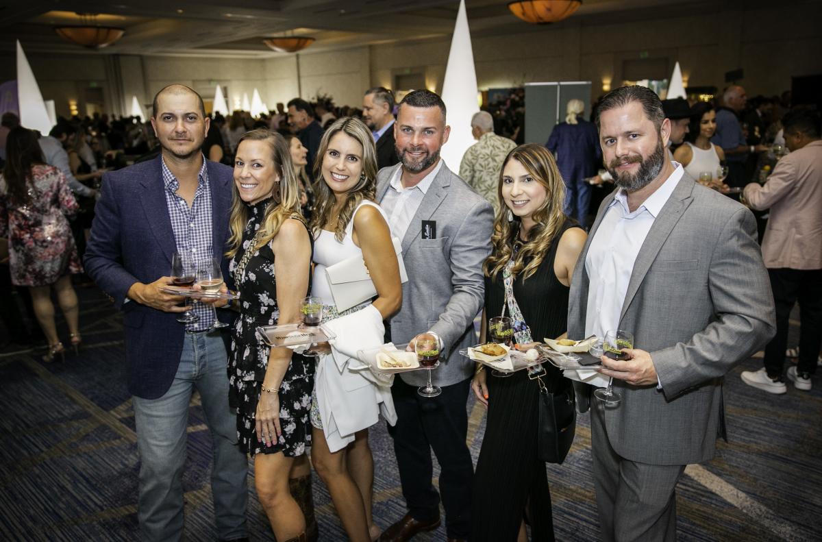 Six people (three men, two women, all in suits or summer dresses) smile at the camera while holding glasses of white and small plates for tastings. Behind them is a room full of people, also mingling and enjoying Wine & Food Week.
