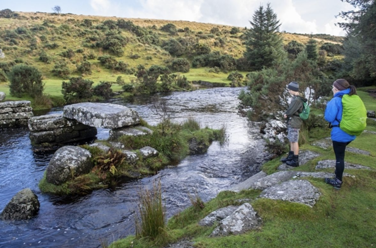 Two hikers with colourful backpacks stand beside a rocky stream in a lush, green landscape, conveying a sense of adventure and tranquillity.