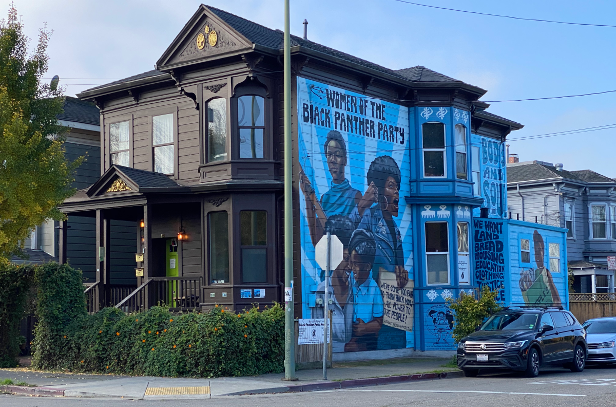 A brown Victorian home stands on a street corner. Colorful  murals depict the Women of the Black Panther Party calling for justice.