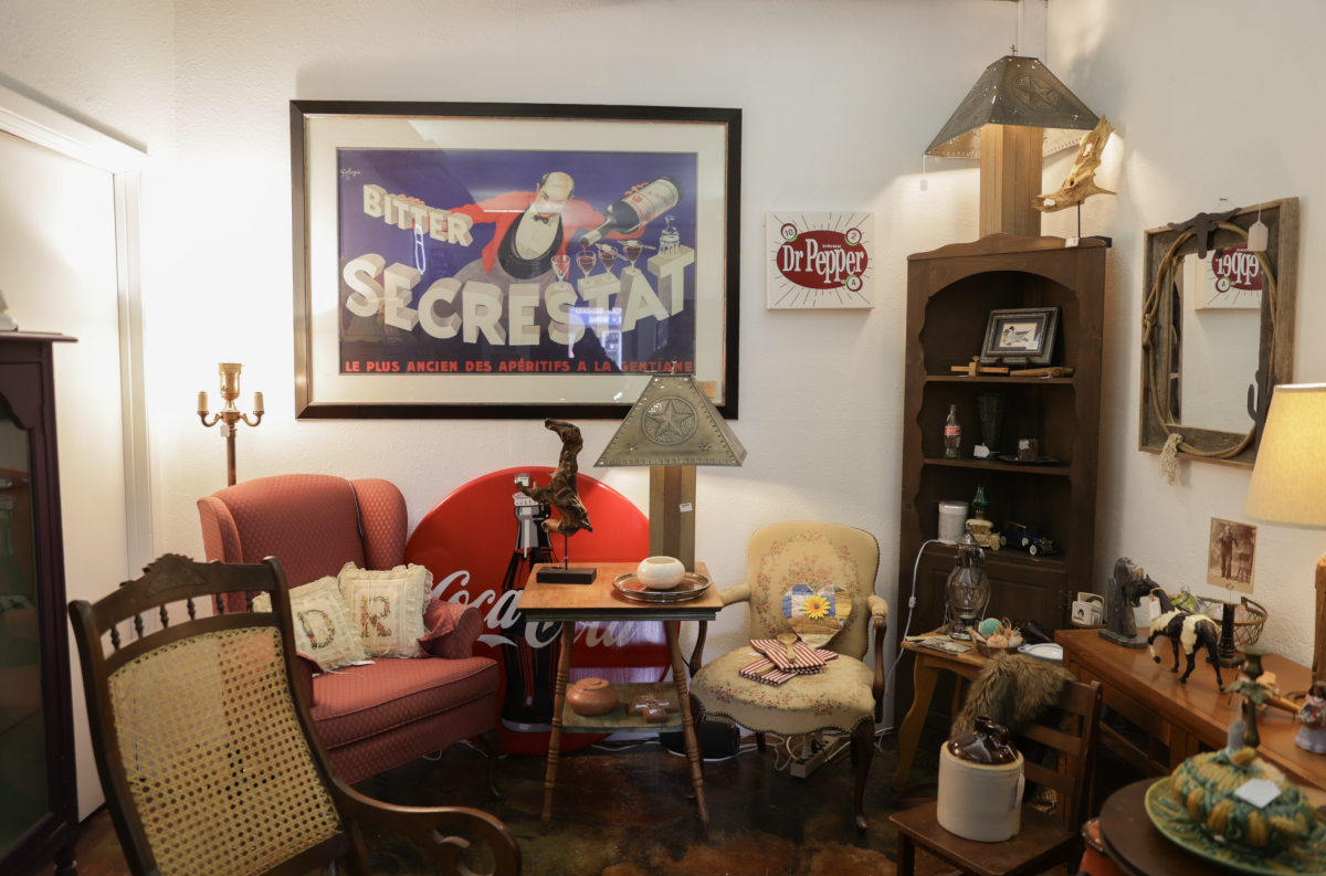 A cozy antique-filled corner at Bluebonnet Hill Furniture and Antiques, featuring a mix of vintage furniture, rustic decor, and nostalgic memorabilia. The space includes a red upholstered armchair with decorative pillows, a wooden rocking chair with a woven seat, and an ornate yellow floral chair. A framed vintage French liquor advertisement hangs above, alongside a Dr. Pepper sign. A Coca-Cola sign leans against the wall, and shelves display small collectibles, books, and rustic trinkets. The warm lighting and eclectic arrangement create a nostalgic and inviting atmosphere.