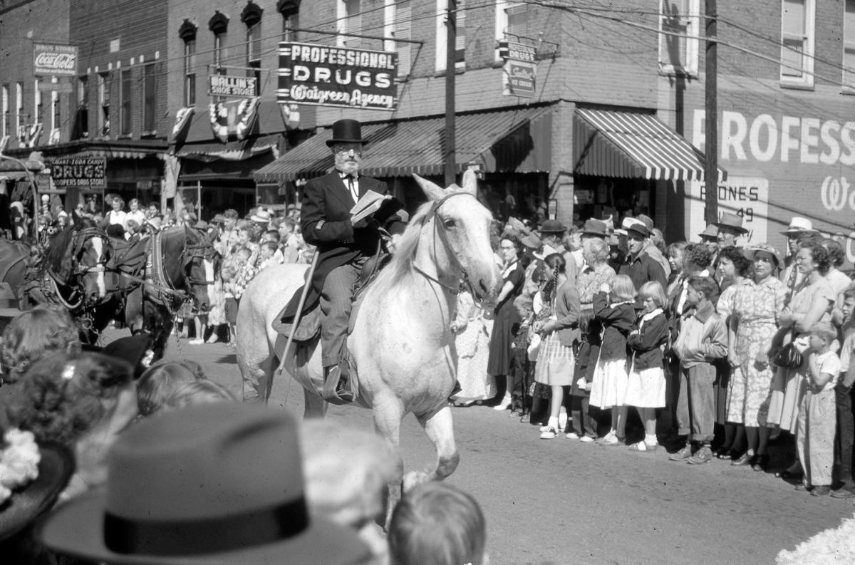 Bearded man in a top hat riding a white horse in the 1951 Jackson County Centennial Parade past crowds and Professional Drugs in Sylva, NC