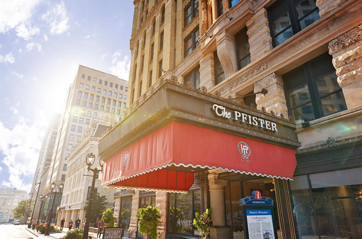 Exterior photo of The Pfister Hotel in downtown Milwaukee on a sunny day. The historic stone building has ornate detailing and a red entrance awning with “The Pfister” signage above it. Sunlight flares in the sky, and the street, sidewalks, and nearby buildings are visible along the block.