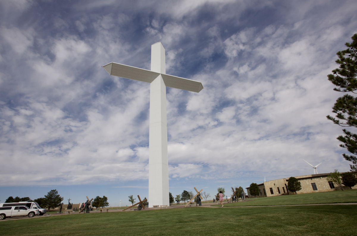 The towering white cross at Groom, Texas rises high into a dramatic sky, surrounded by green lawns and bronze sculptures depicting the Stations of the Cross. Visitors walk the peaceful grounds, while nearby buildings and wind turbines dot the Panhandle landscape.