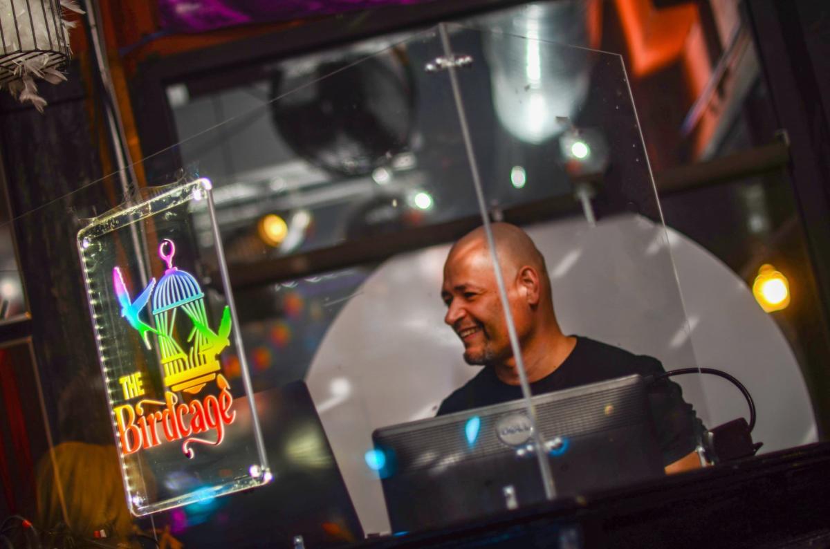 Image is of a male DJ behind the glass which says "The Birdcage" and has their logo.