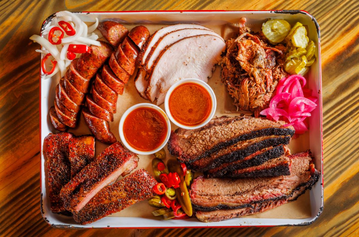 Flat lay image of a metal tray with different kinds of smoked meats, sides and barbecue sauces.