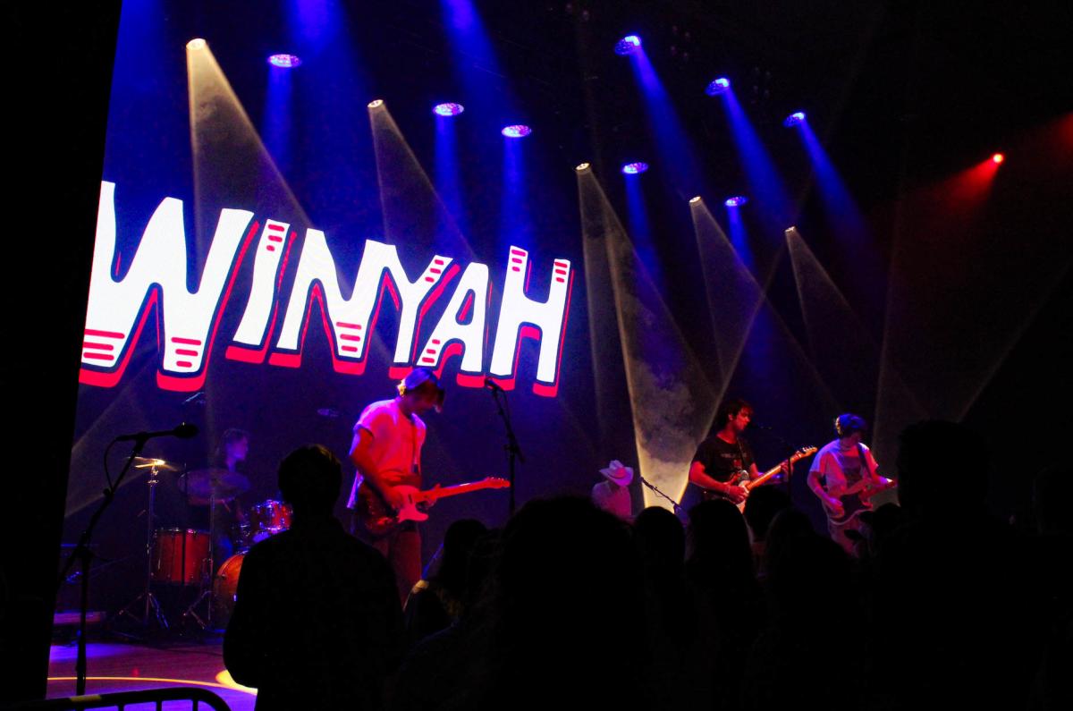 A live music performance featuring a band on stage with vibrant lighting. The backdrop displays the word "WINYAH" in bold letters.