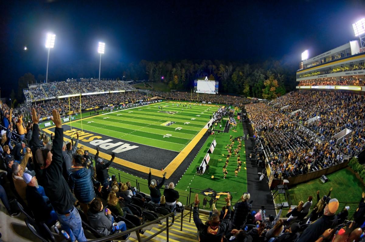 A football stadium filled with fans in black and gold cheer on the Appalachian State Mountaineers during a night time home game.