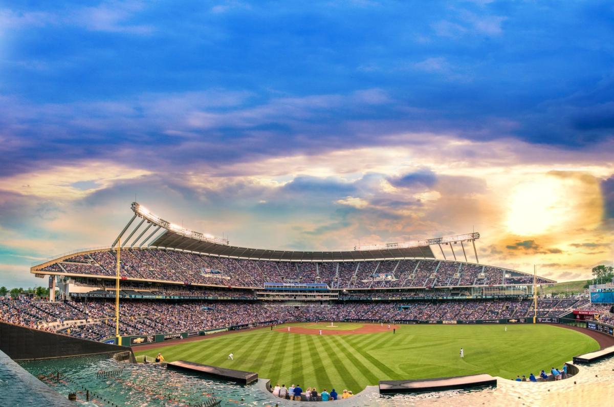 The green baseball field is in the foreground while the sun sets behind Kauffman Stadium