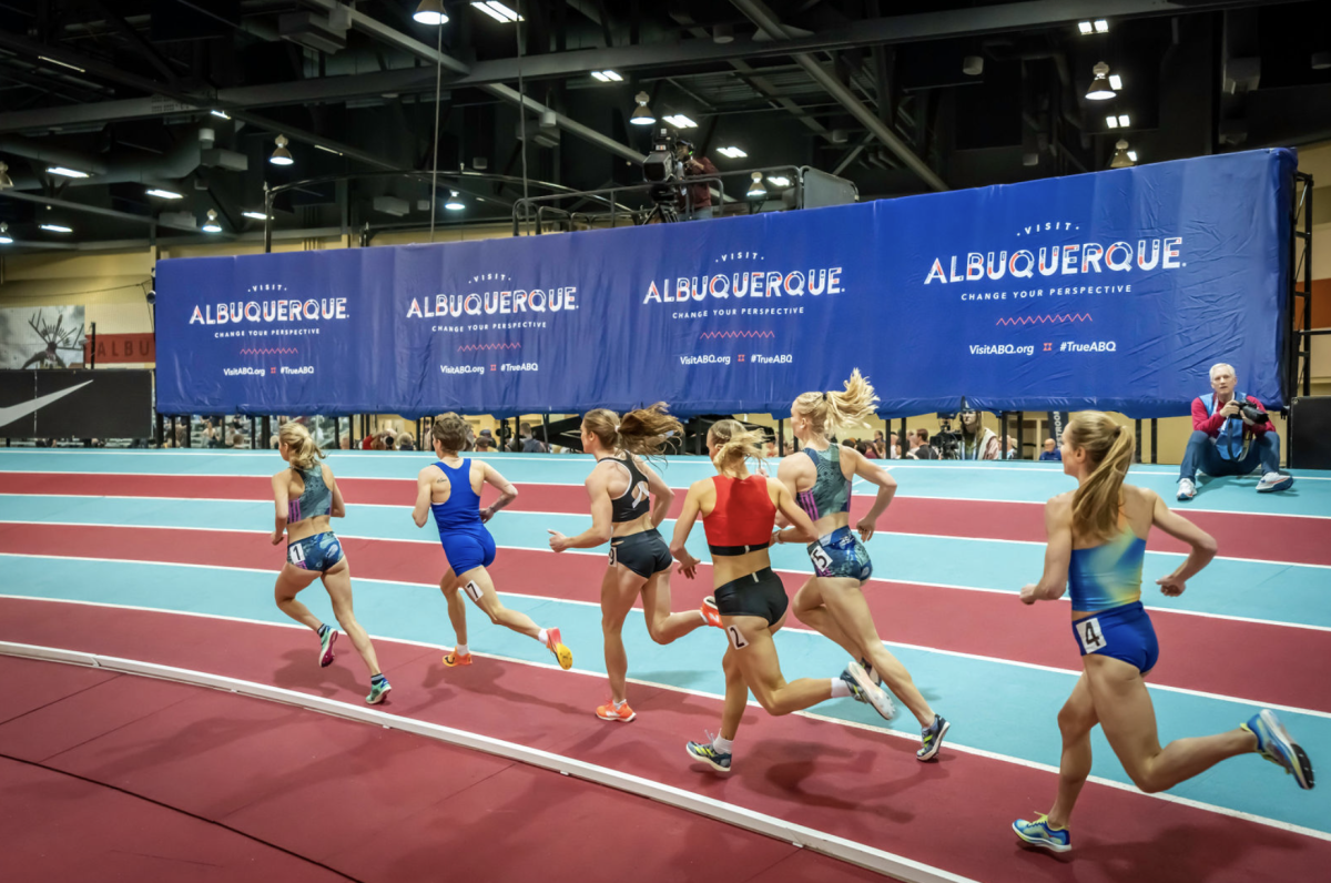 Runners run on a track at the Albuquerque Convention Center with a crash pad in the background that has the Visit Albuquerque logo on it at the 2024 USATF Indoor Championships