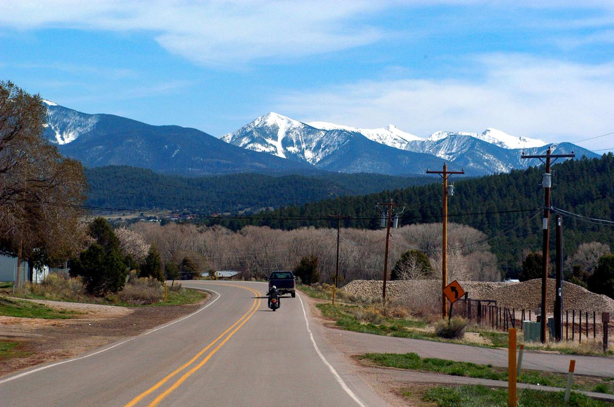 A scenic road leading towards snow-capped mountains, with a vehicle and motorcycle traveling along it. Forested hills and open fields line the road under a partly cloudy sky.