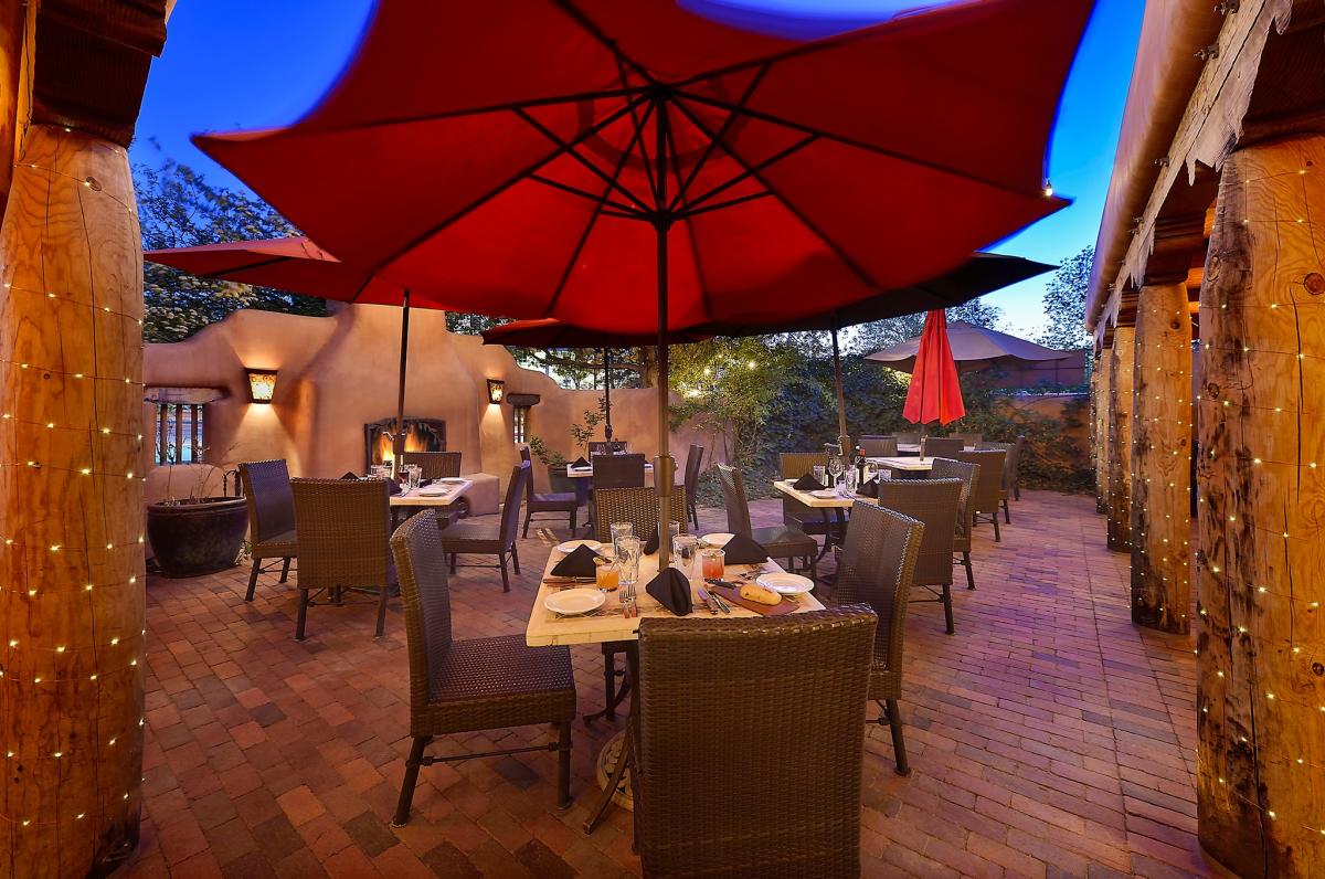 Rustic outdoor restaurant patio with warm lighting, brick flooring, and red umbrellas. Wicker chairs surround tables set for dining.