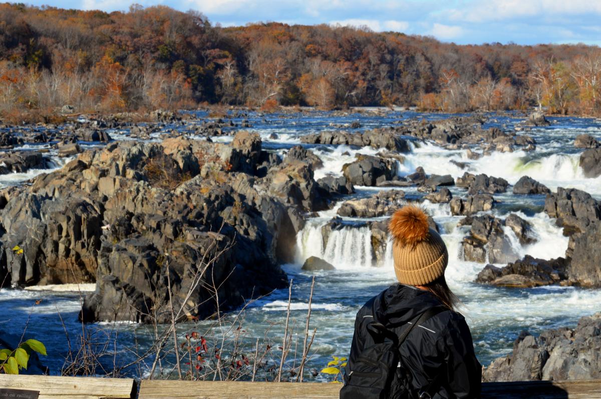 A person in a brown beanie gazes at a cascading waterfall surrounded by rocky terrain and autumn foliage, evoking natural beauty.