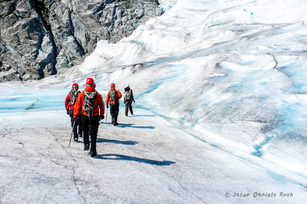 A group of people in red jackets and helmets touring the icy blue Mendenhall Glacier.