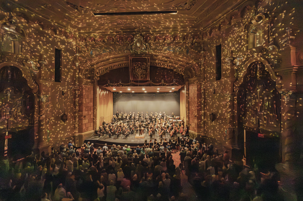 Interior of the Frauenthal Center in downtown Muskegon with a West Michigan Symphony performing on stage, warmly illuminated by golden lights with a captivated audience in attendance.