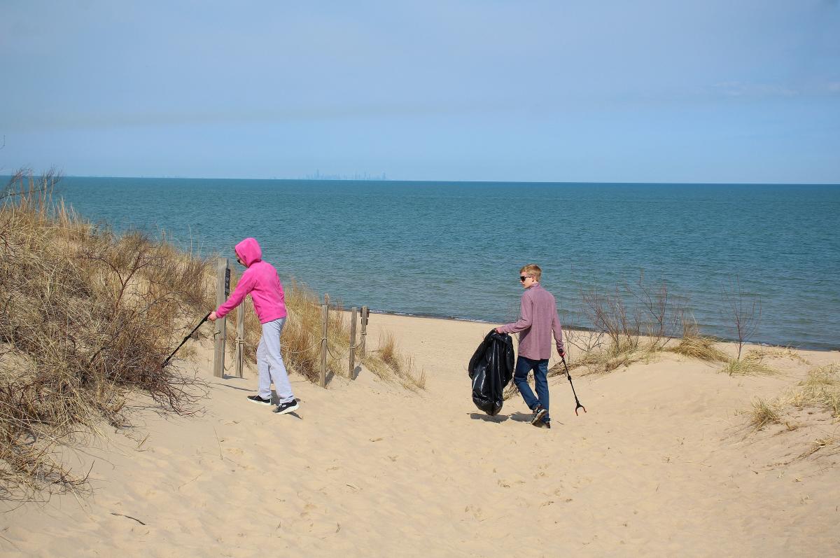 Two people walk along a sandy lakeshore with trash grabbers and a black garbage bag