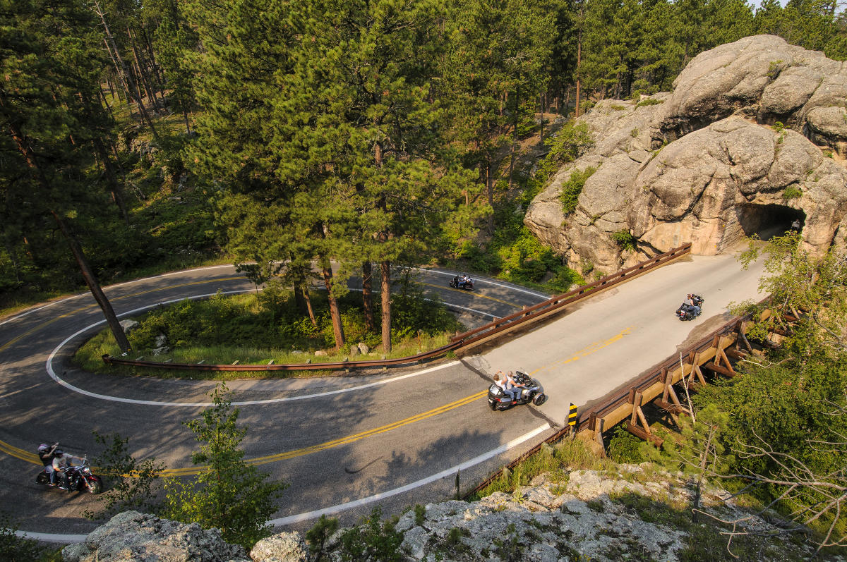 From a high angle, three motorcycles ride around the iconic pigtail turns on Iron Mountain Road approaching a tunnel on the upper right side of the photo.