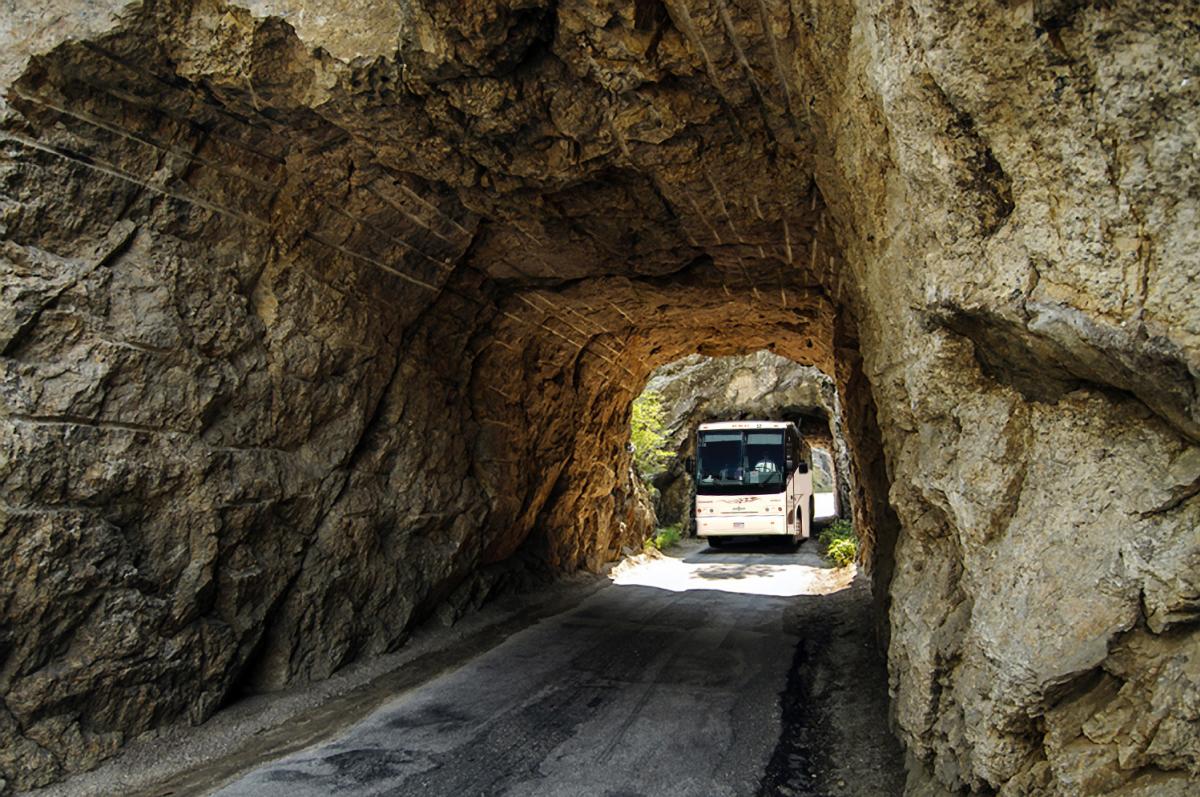 A white RV carefully navigates a narrow, rocky tunnel carved from stone. Sunlight highlights parts of the rough-textured walls, creating a tense yet awe-inspiring scene.